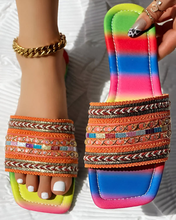 Une femme porte des sandales plates avec des perles et des broderies colorées. Les sandales ont une semelle intérieure orange et une semelle extérieure multicolore. La femme porte également un bracelet de cheville doré.