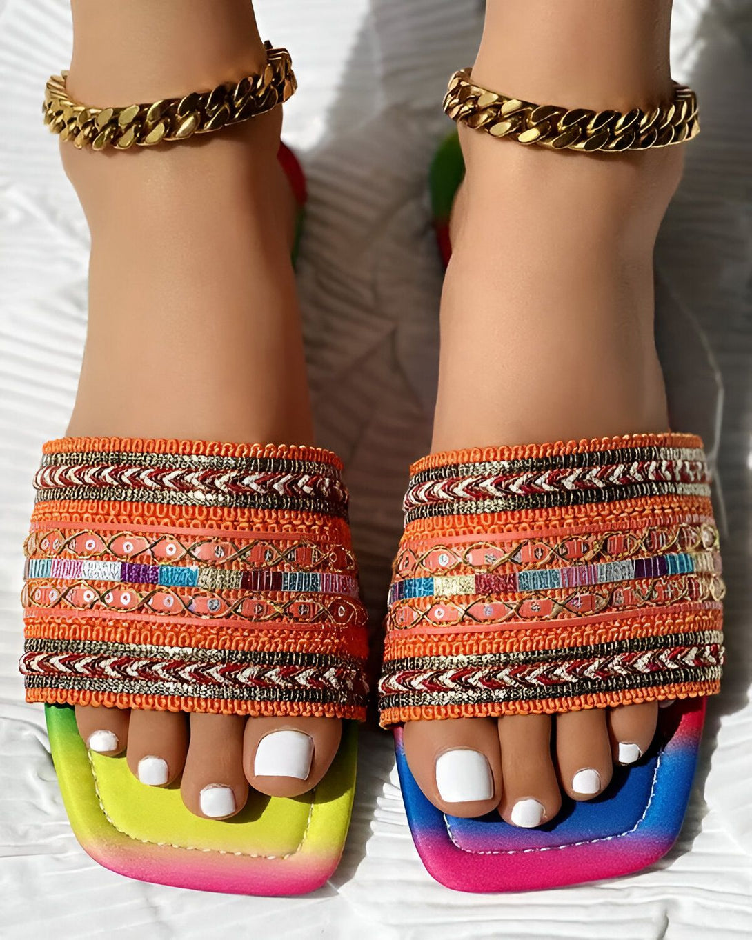 Une femme porte des sandales plates multicolores avec des perles et des broderies. Les sandales ont une semelle intérieure orange et une semelle extérieure verte, jaune, orange et rose. La femme porte également des bracelets de cheville en or.