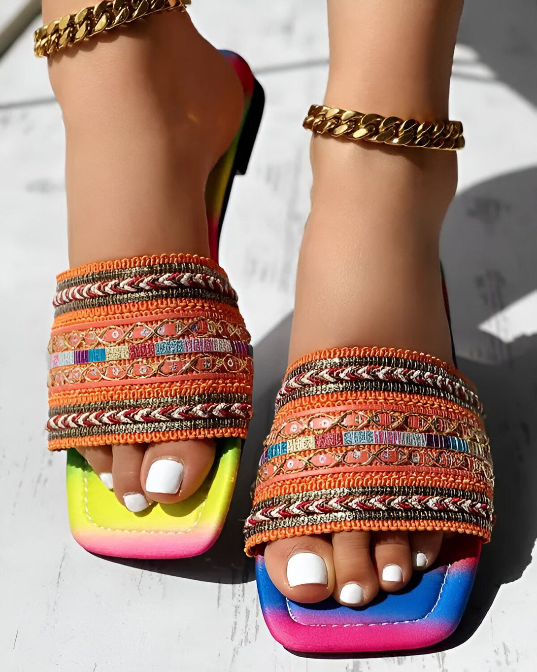 Une femme porte des sandales plates multicolores avec des motifs ethniques. Les sandales sont ouvertes au niveau du talon et ont une bride qui passe entre les orteils. La femme a les ongles des pieds peints en blanc. Elle porte également un bracelet de cheville doré.