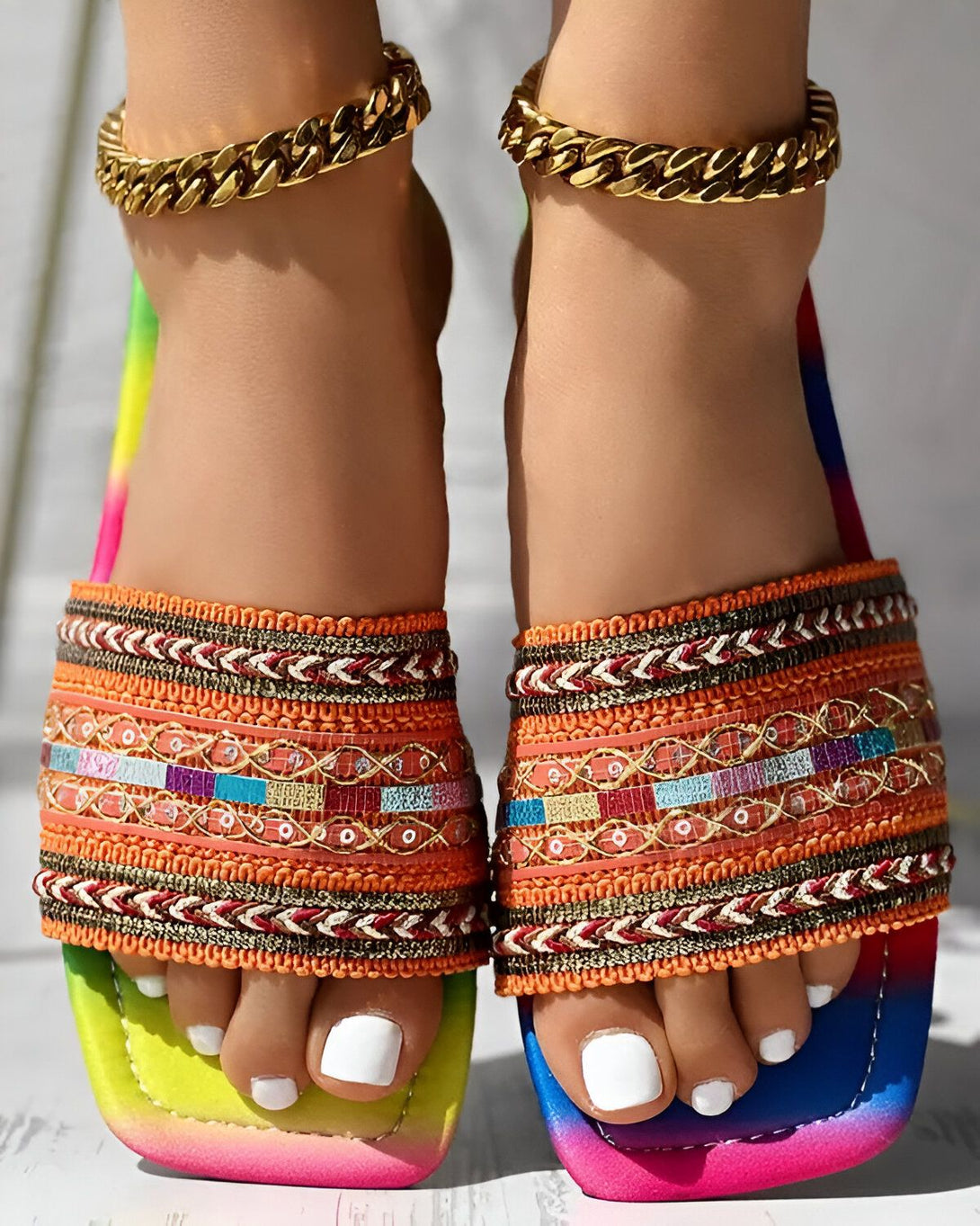 Une femme porte des sandales plates multicolores avec des perles et des broderies. Les sandales ont une semelle intérieure blanche et une semelle extérieure multicolore. La femme a les ongles des pieds peints en blanc et porte des bracelets de cheville en or.