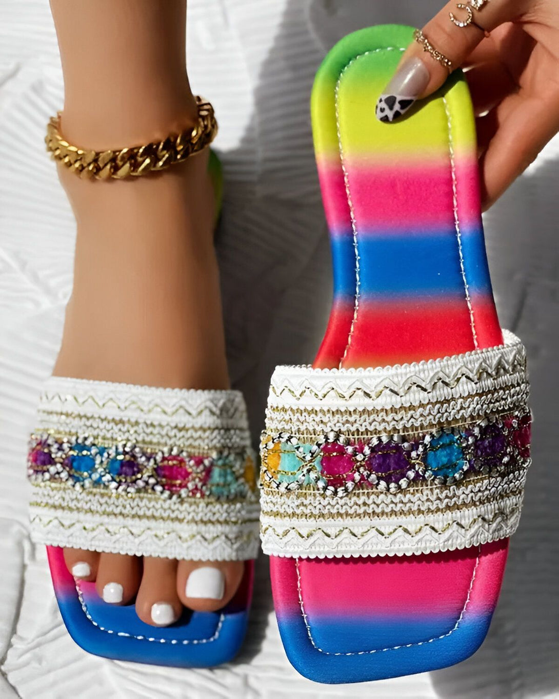 Une femme porte des sandales plates à enfiler. Les sandales sont blanches avec des perles colorées et des détails dorés. La femme a les ongles des pieds peints en blanc et porte un bracelet de cheville doré.