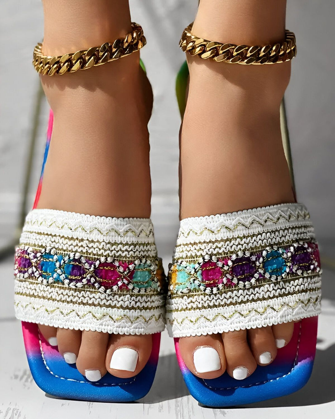 Une femme porte des sandales plates avec des brides colorées et des détails brodés. Les ongles des pieds de la femme sont peints en blanc.
