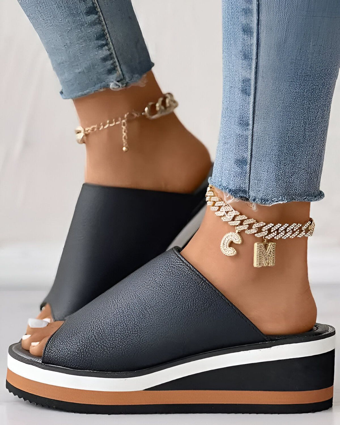 Une femme porte des mules noires à plateforme avec un bijou de cheville doré avec un pendentif en forme de lettre "M". Elle porte également un jean bleu.