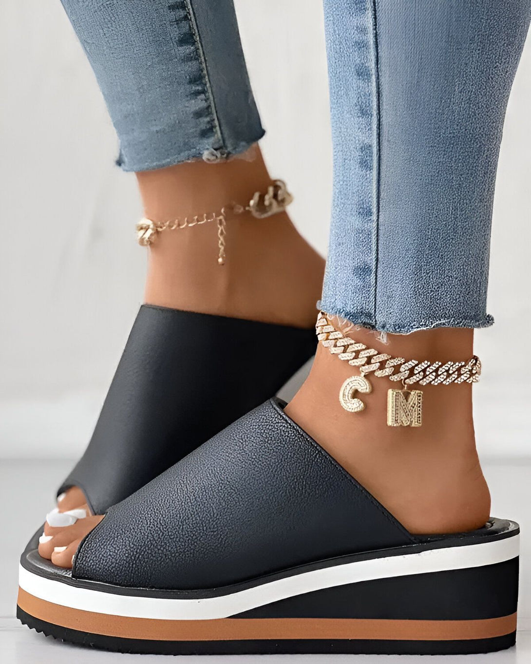 Une femme porte des sandales noires à talons compensés. Les sandales sont ouvertes au niveau des orteils et ont une bride qui passe sur le dessus du pied. La femme porte également un jean bleu et des bijoux dorés.