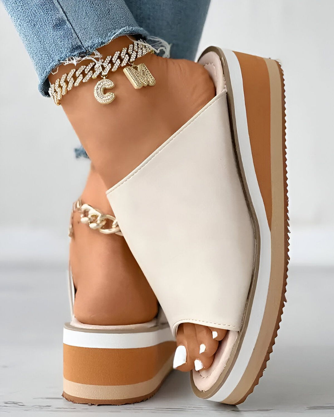 Une femme porte des sandales compensées beiges et blanches. Les sandales sont ouvertes au niveau des orteils et ont une bride qui passe sur le dessus du pied. La femme porte également un jean bleu et un bracelet de cheville doré.