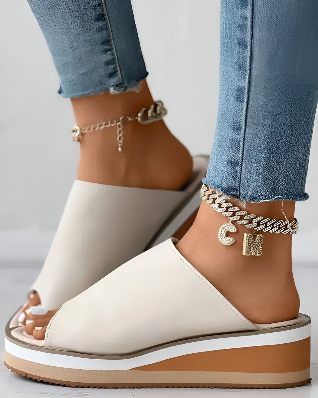 Une femme porte des sandales blanches à talons compensés. Les sandales sont ouvertes sur le devant et ont une bride qui passe sur le dessus du pied. La femme porte également un jean bleu et des bijoux dorés.