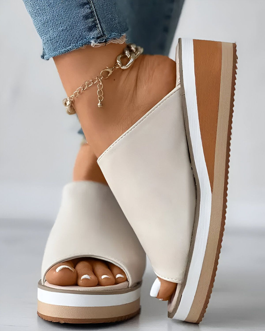 Une femme porte des mules compensées blanches. Les mules ont une bride sur le dessus et sont ouvertes au niveau du talon. La femme porte un jean bleu et un bracelet de cheville doré.