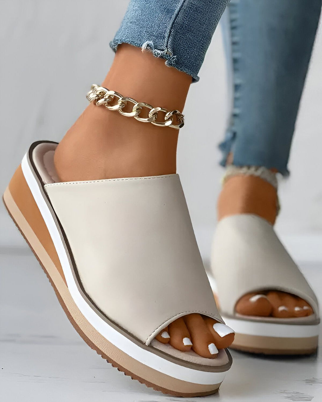 Une femme porte des sandales beiges à talons compensés. Les sandales sont ouvertes au niveau des orteils et ont une bride qui passe sur le dessus du pied. La femme porte également un jean bleu et un bracelet de cheville doré.