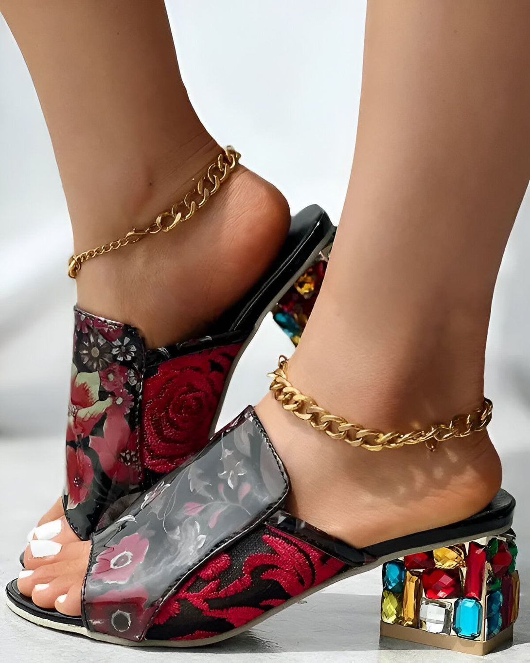 Une femme porte des mules à talons hauts à bout ouvert. Les mules sont noires avec un motif floral coloré. Le talon est recouvert de pierres précieuses multicolores. La femme a les ongles des pieds peints en blanc. Elle porte également une chaîne dorée à la cheville.