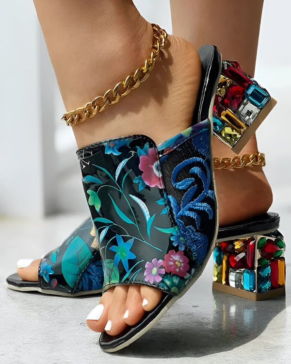 Une femme porte des mules à talons hauts. Les mules sont noires et à motifs floraux. Le talon est transparent et coloré. La femme porte également une chaîne dorée autour de la cheville.