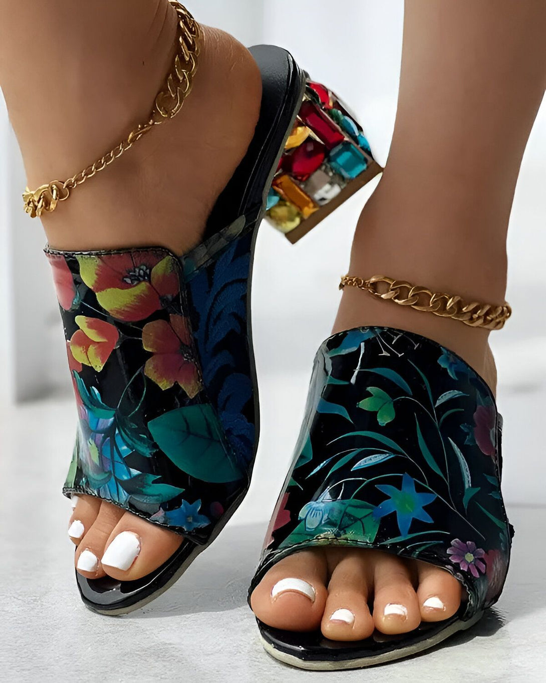 Une femme porte des mules à talons hauts à fleurs. Les mules sont noires avec un imprimé floral coloré. Les talons sont transparents et ont une forme géométrique. La femme a les ongles des pieds blancs et porte des bijoux de cheville en or.
