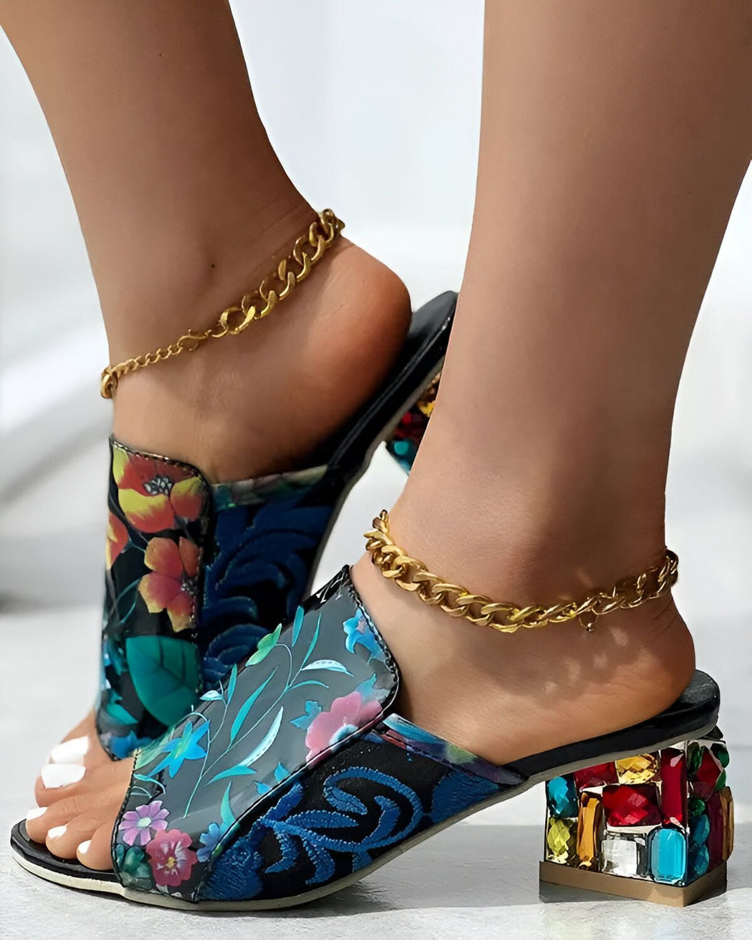 Une femme porte des mules à talons hauts à bout ouvert. Les mules sont de couleur bleu marine et ont un motif floral coloré. Le talon est transparent et incrusté de pierres précieuses colorées. La femme porte également une chaîne dorée autour de la cheville.
