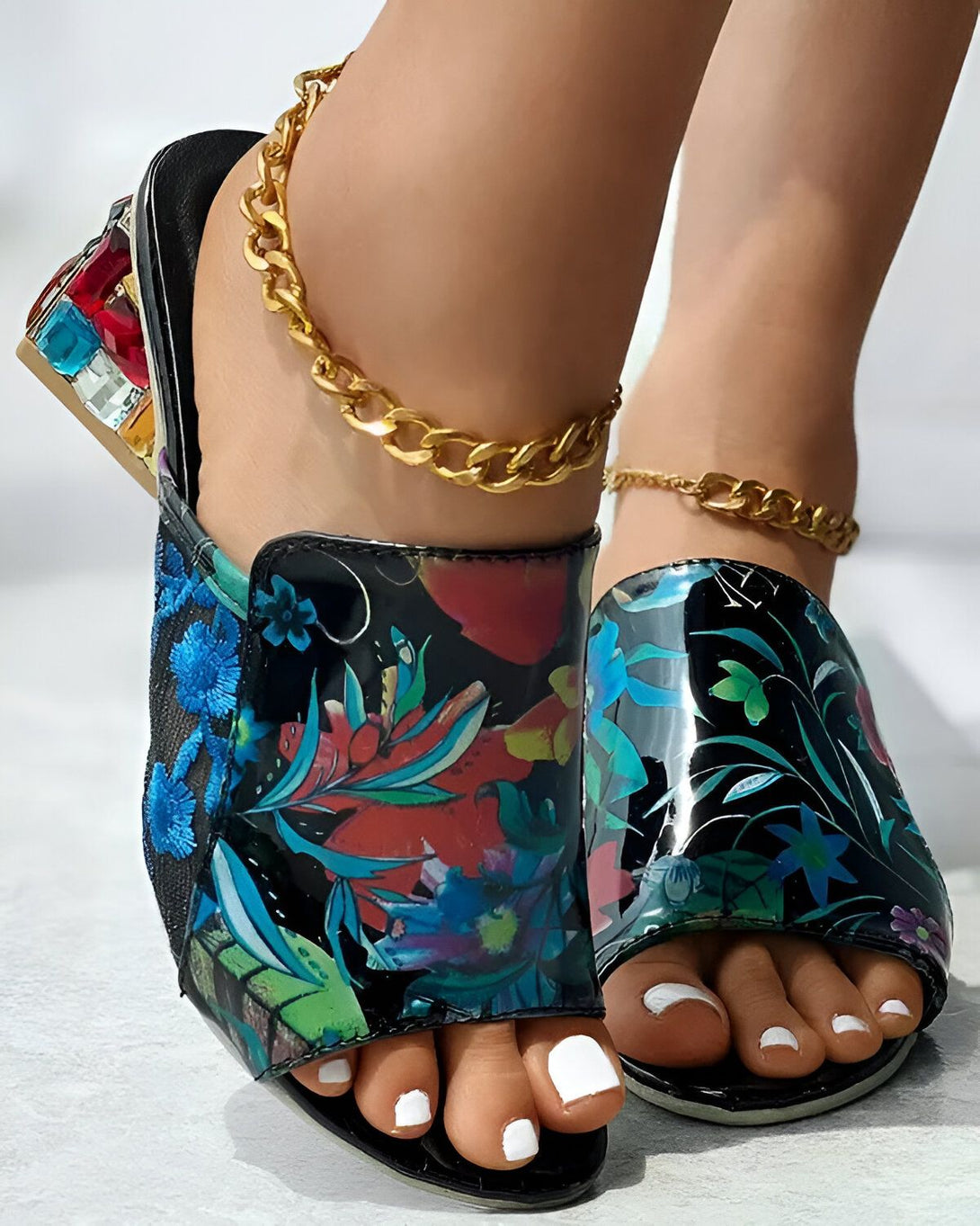 Une femme porte des mules à talons hauts à bout ouvert. Les mules sont noires et à motifs floraux colorés. La femme a les ongles des pieds blancs et porte des bijoux de cheville en or.