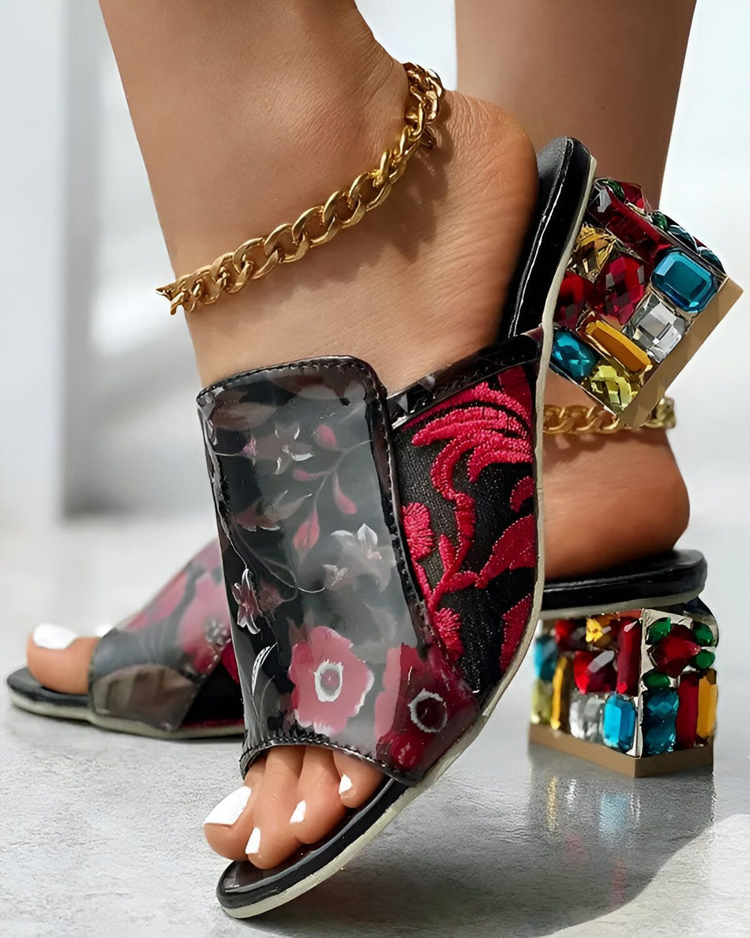Une femme porte des mules à talons hauts noirs avec des fleurs rouges brodées et des pierres précieuses multicolores sur le talon.