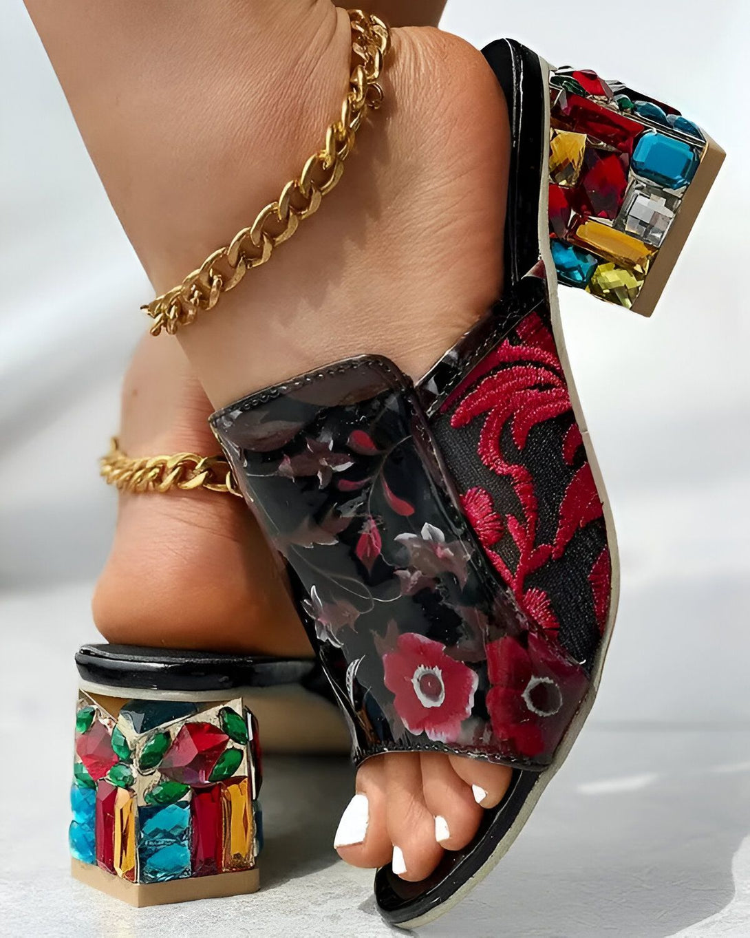 Une femme porte des mules à talons hauts noires et rouges. Les mules sont ornées de broderies florales et de pierres précieuses colorées. La femme porte également une chaîne dorée autour de la cheville.