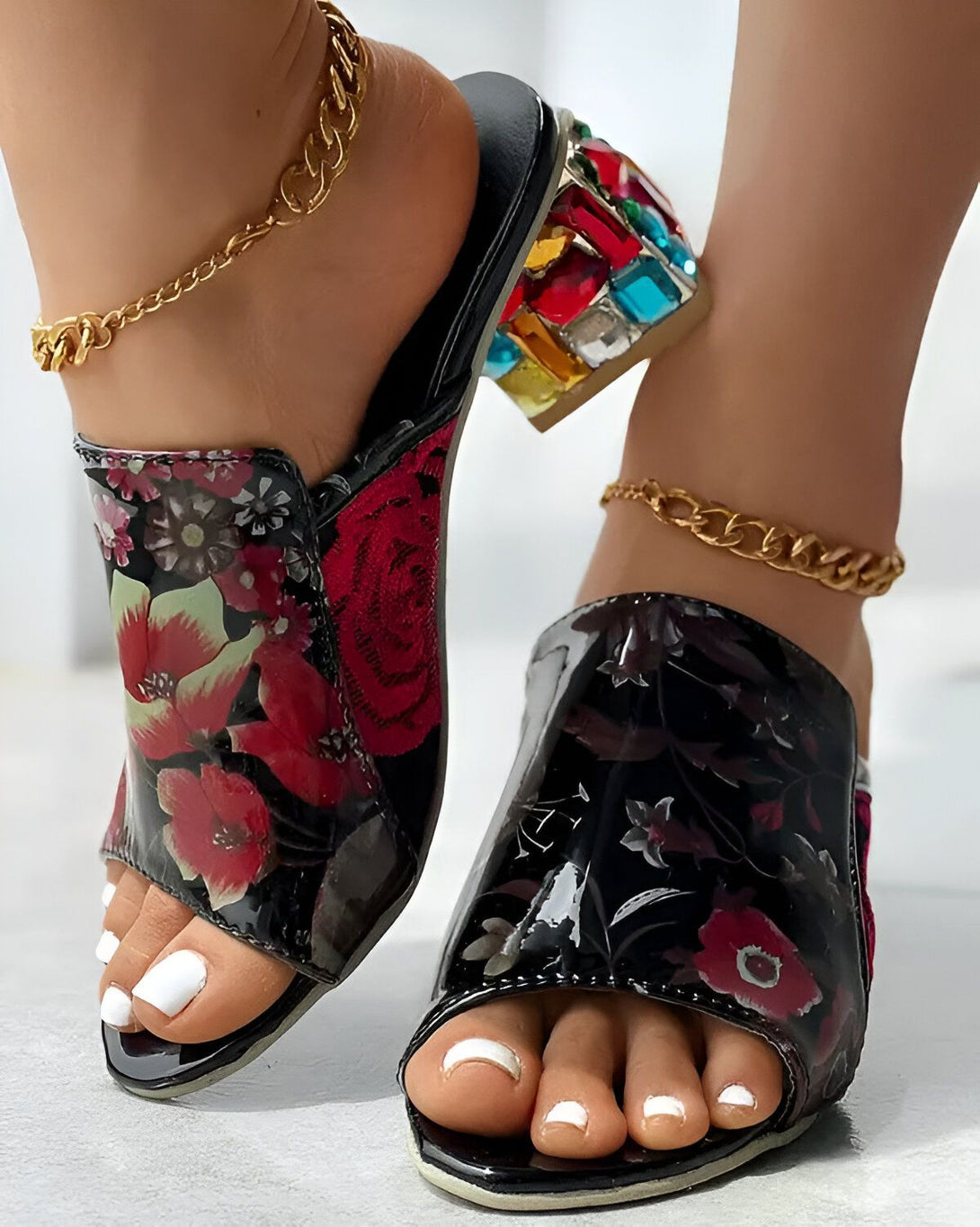 Une femme porte des mules à talons hauts noires et rouges à fleurs. Les mules sont ornées de cristaux multicolores. La femme a les ongles des pieds peints en blanc. Elle porte également une chaîne dorée à la cheville.