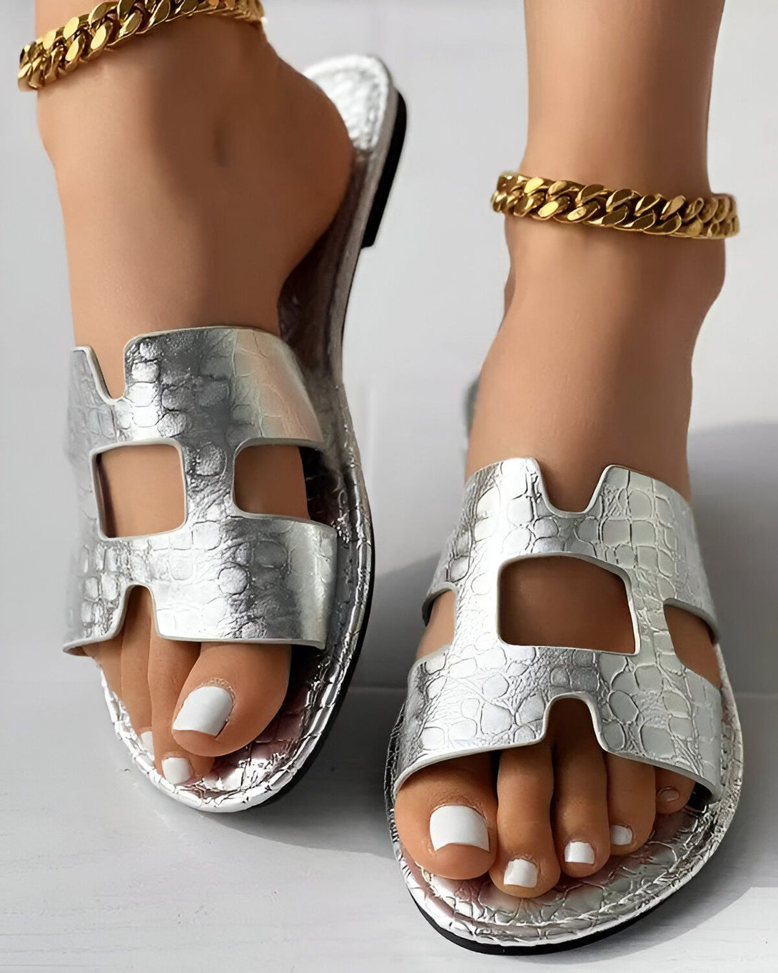 Une paire de sandales plates en cuir argenté métallisé avec une bride en H sur le dessus. Les sandales sont portées par une femme avec un pantalon beige et un haut blanc.