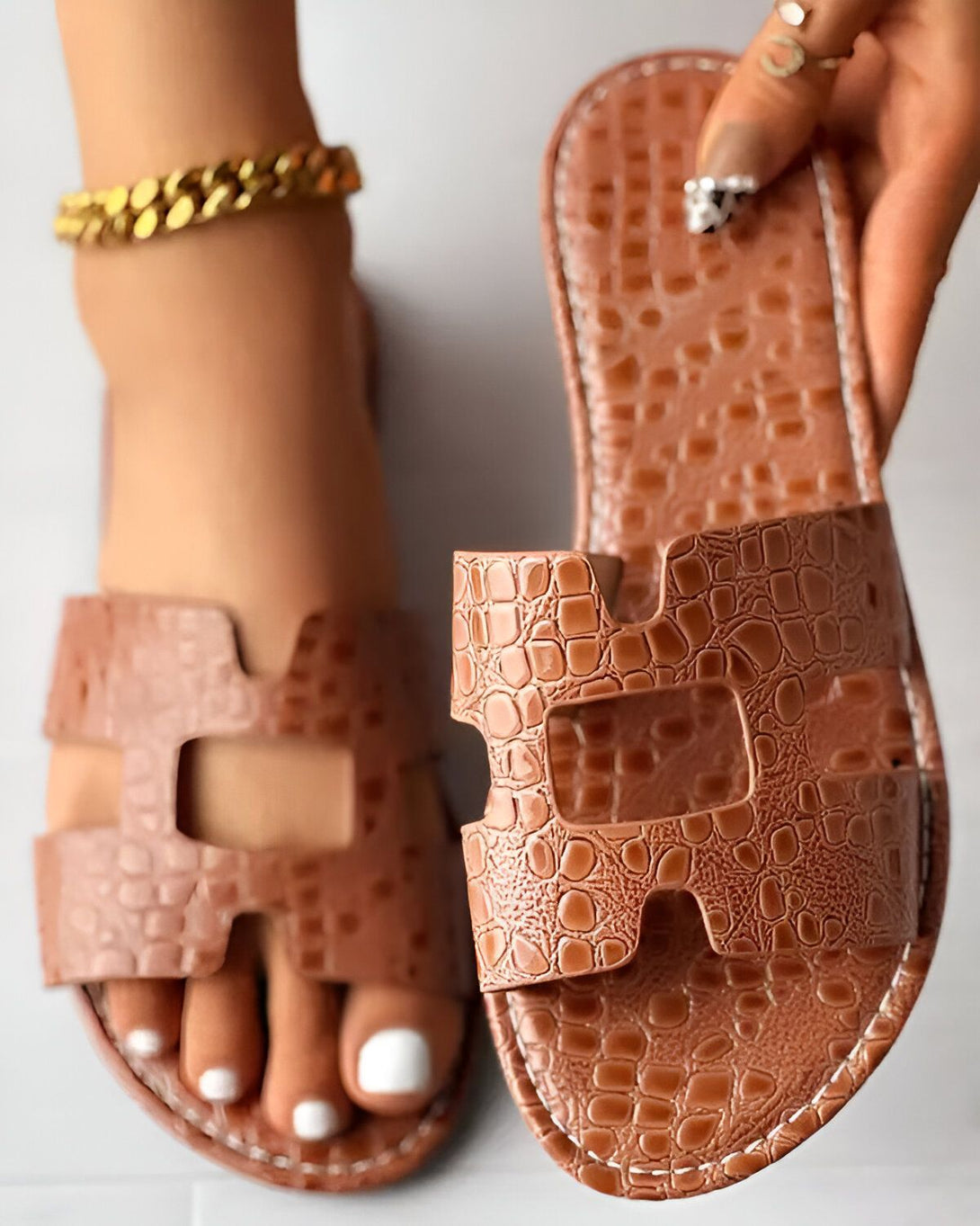 Une paire de sandales marron en cuir avec un motif croco. Les sandales sont portées par une femme qui a une chaîne en or à la cheville. La femme a les ongles des pieds peints en blanc.