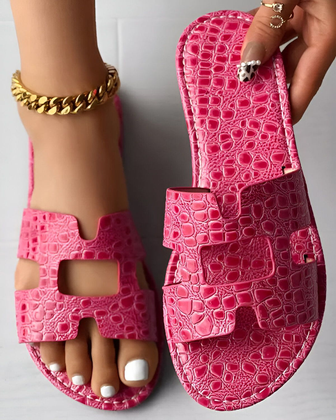 Une femme porte des sandales roses à talons hauts. Les sandales sont en cuir et ont un design croisé sur le dessus. La femme a les ongles des pieds peints en blanc et porte un bracelet de cheville en or.