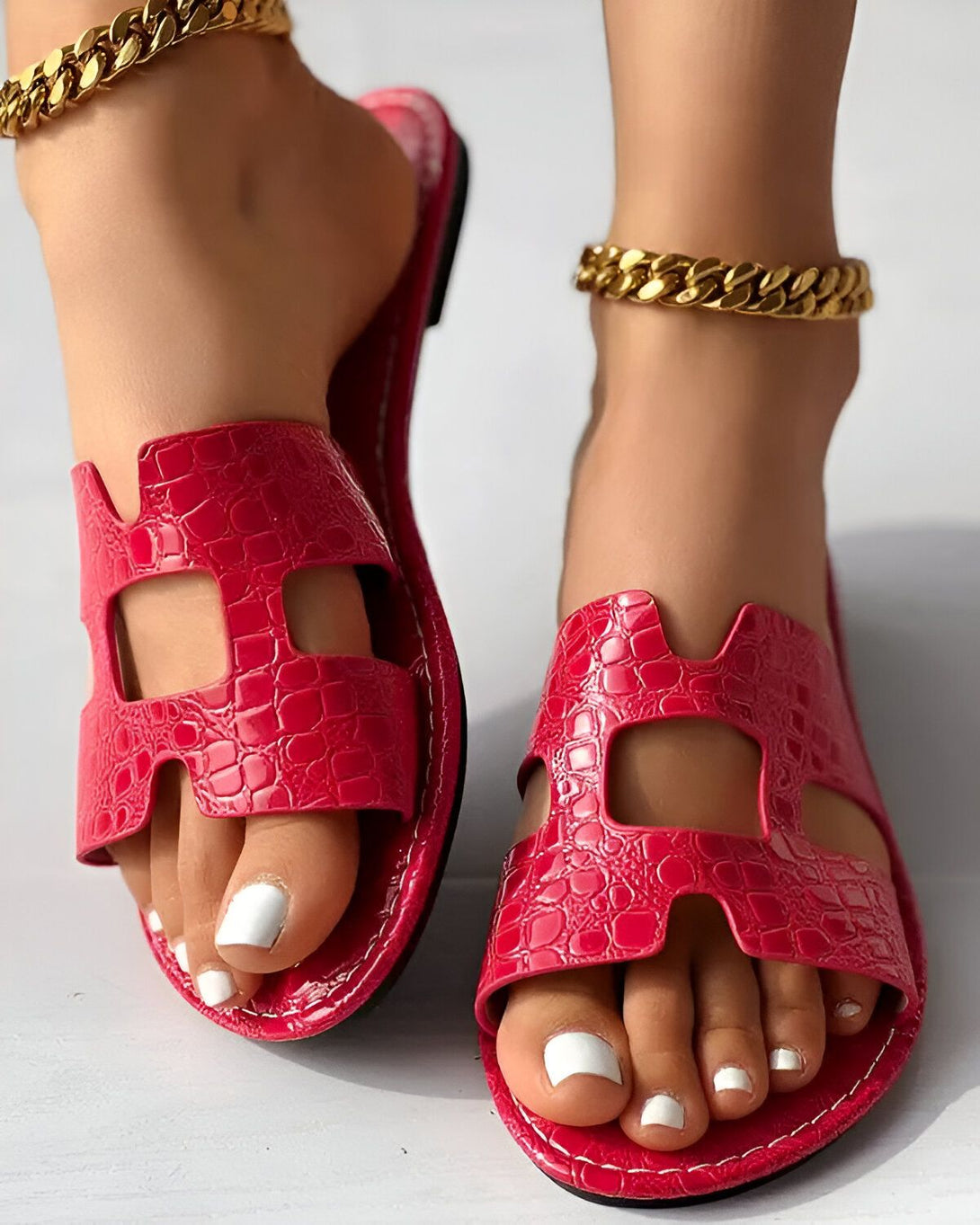 Une femme porte des sandales rouges à talons hauts. Les sandales sont en cuir et ont un design croisé sur le dessus. La femme a les ongles des pieds peints en blanc et porte des bracelets de cheville en or.