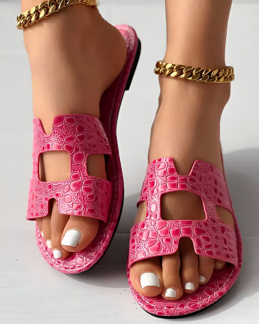Une femme porte des sandales roses avec des chaînes dorées aux chevilles. Les sandales sont en cuir et ont un design croisé sur le dessus. La femme a les ongles des pieds peints en blanc.