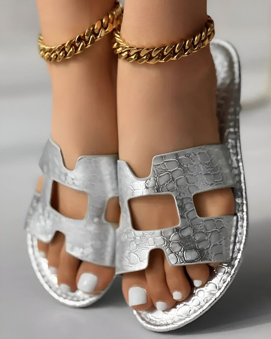 Une paire de sandales argentées à talons hauts avec des brides en forme de H. Les sandales sont portées par un mannequin avec des chevilles fines et des ongles blancs. Le mannequin porte également des bracelets de cheville en or.