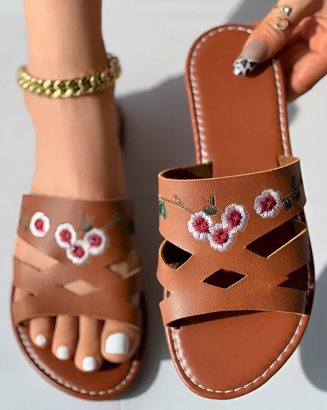 Une paire de sandales plates en cuir marron avec des brides croisées sur le dessus. Les brides sont brodées de fleurs roses et blanches. Les sandales sont portées par une femme qui a les ongles des pieds peints en blanc. Elle porte également un bracelet de cheville en or.