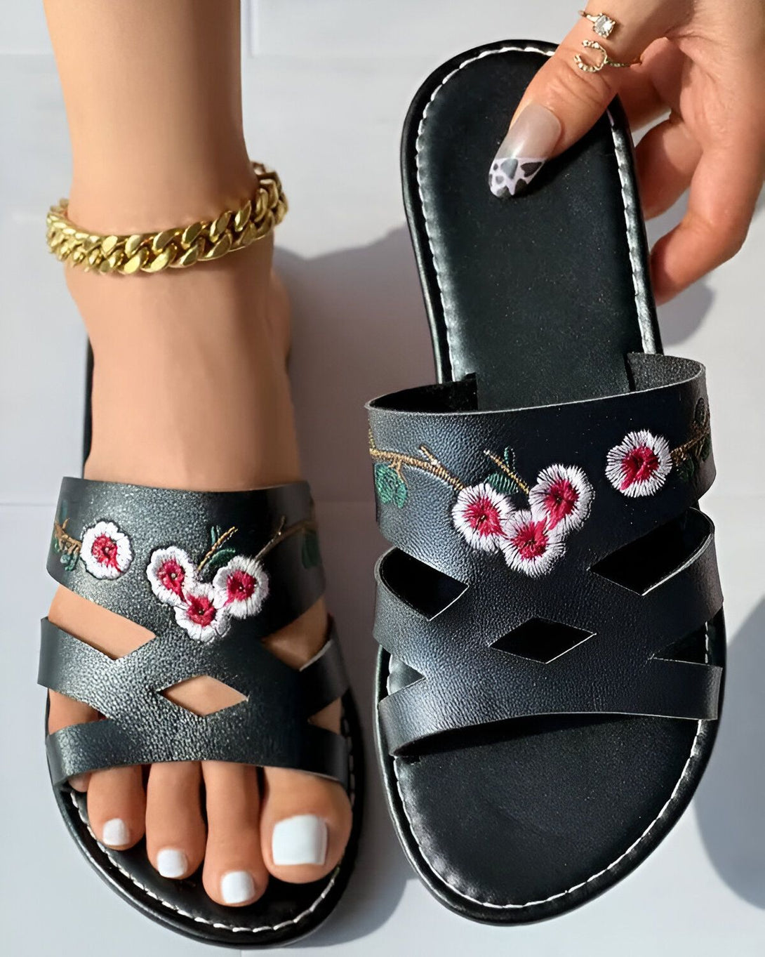 Une image de sandales plates noires avec des fleurs brodées dessus. Les sandales sont portées par une femme qui a une chaîne en or à la cheville et un vernis à ongles rose et noir.