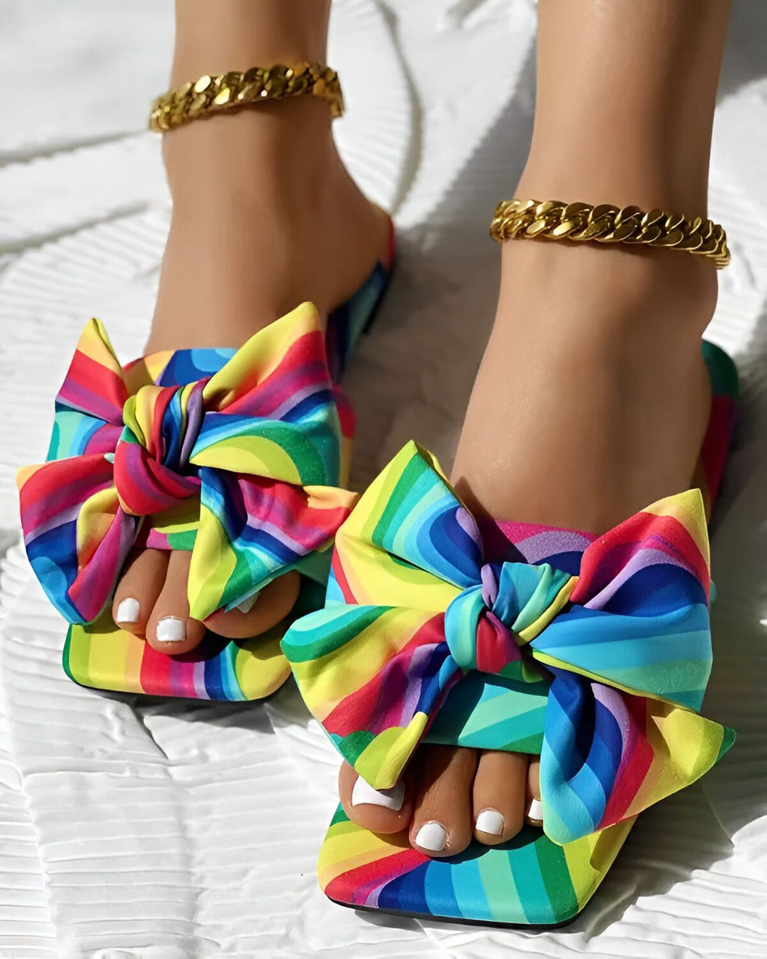 Une femme porte des sandales à talons hauts avec un nœud coloré. Les sandales sont arc-en-ciel et le nœud est fait d'un tissu à motifs. La femme a les ongles des pieds peints en blanc et porte des bijoux en or.