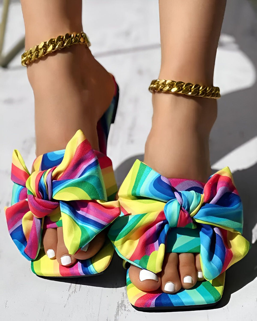 Une femme porte des sandales à talons hauts avec un nœud sur le devant. Les sandales sont multicolores et ont un motif arc-en-ciel. La femme a les ongles des pieds peints en blanc et porte des bijoux en or.