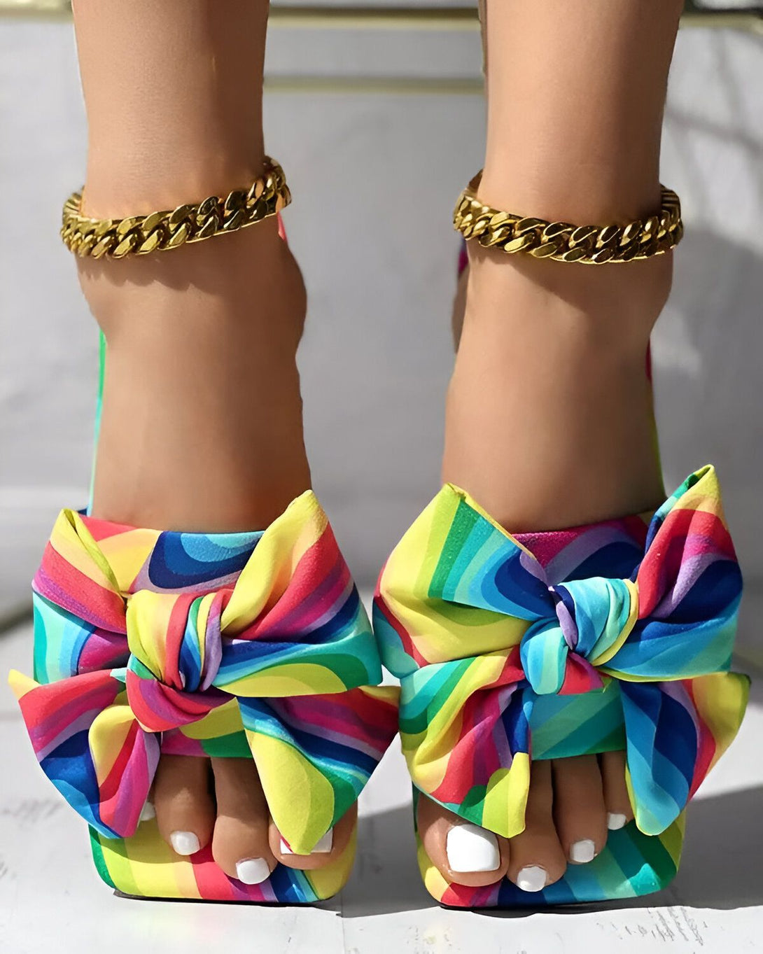 Une femme porte des sandales à talons hauts avec un nœud en tissu arc-en-ciel sur le devant. Elle porte également des bracelets de cheville en chaîne dorée.