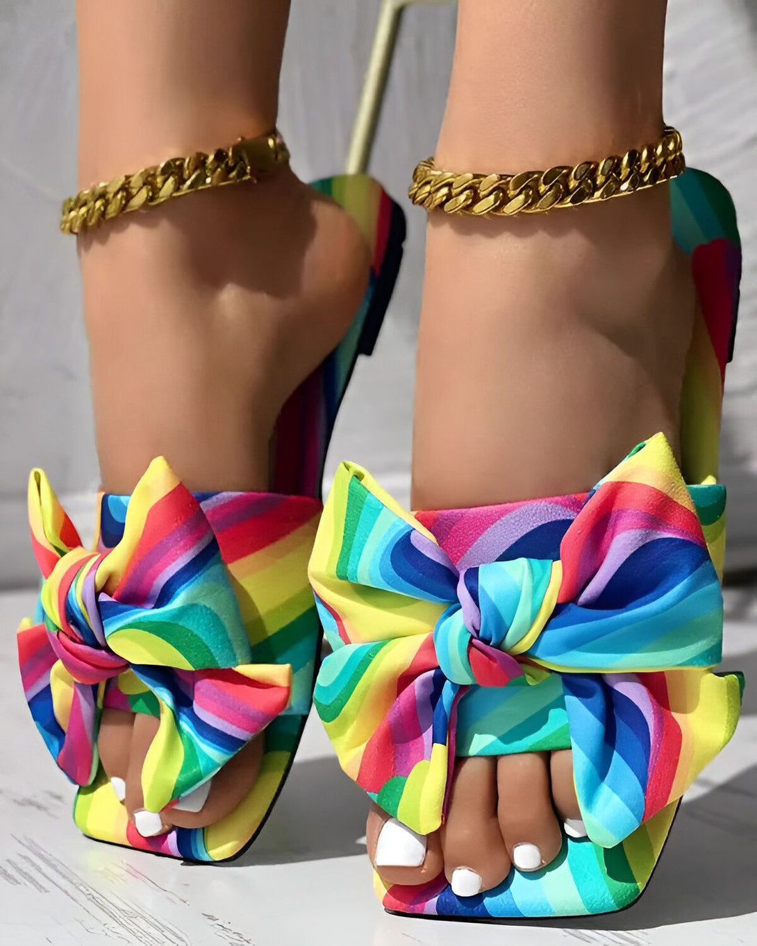 Une femme porte des sandales à talons hauts avec un nœud sur le devant. Les sandales sont multicolores et ont un motif arc-en-ciel. La femme a les ongles des pieds peints en blanc et porte des bijoux de cheville en or.
