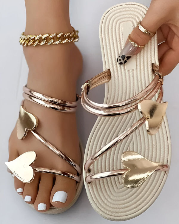 Une femme porte des sandales blanches et dorées. Les sandales ont des cœurs en métal doré sur le dessus. La femme a les ongles des pieds peints en blanc et porte un bracelet de cheville en or.