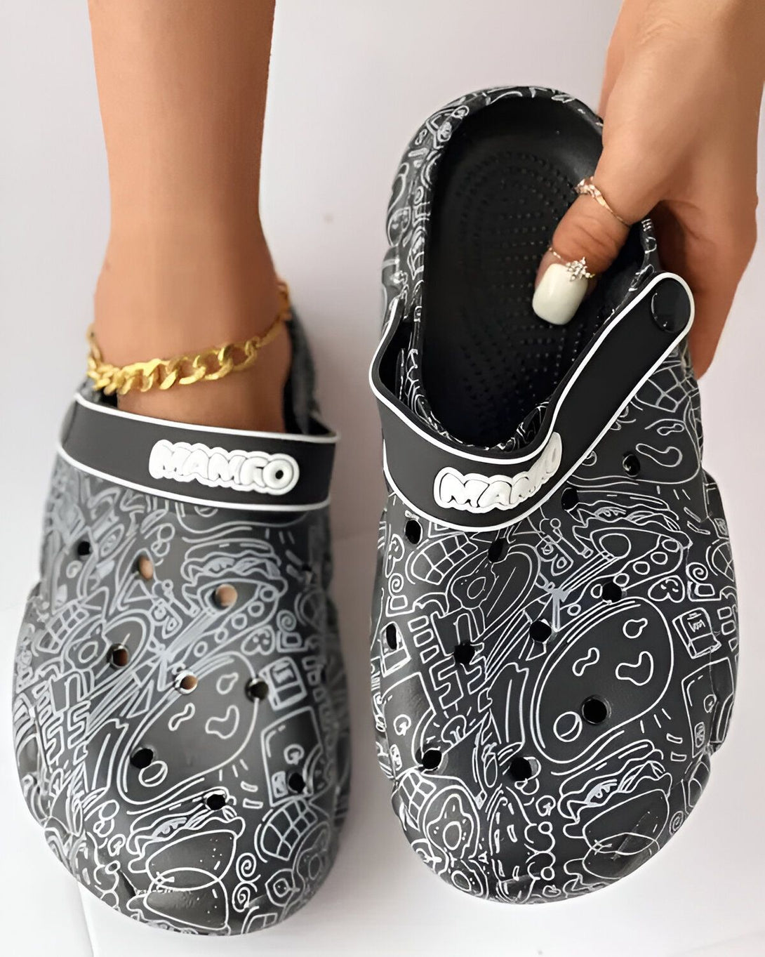 Une paire de sabots noirs et blancs avec des motifs de dessins animés. Les sabots sont portés par une femme qui a une chaîne en or à la cheville.