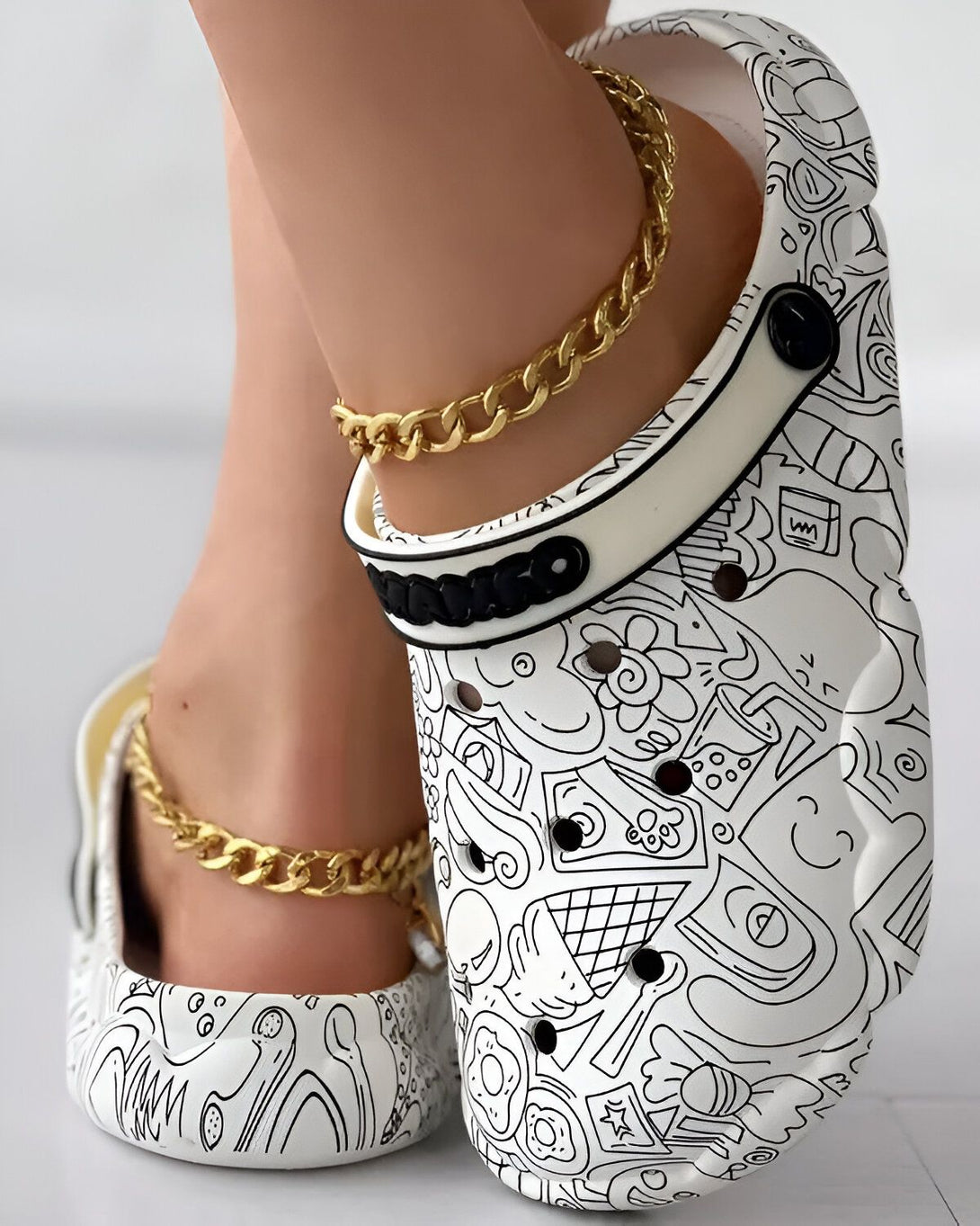 Une femme porte des sandales blanches à motifs noirs et une chaîne de cheville en or.