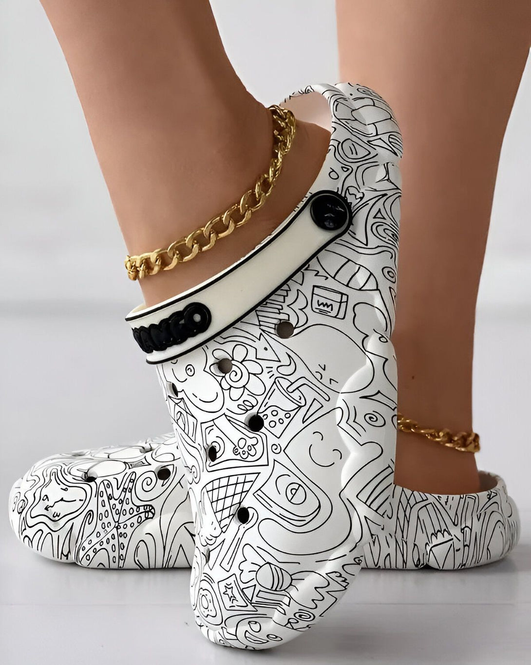Une personne porte des chaussures blanches à motifs noirs avec des chaînes dorées aux chevilles.