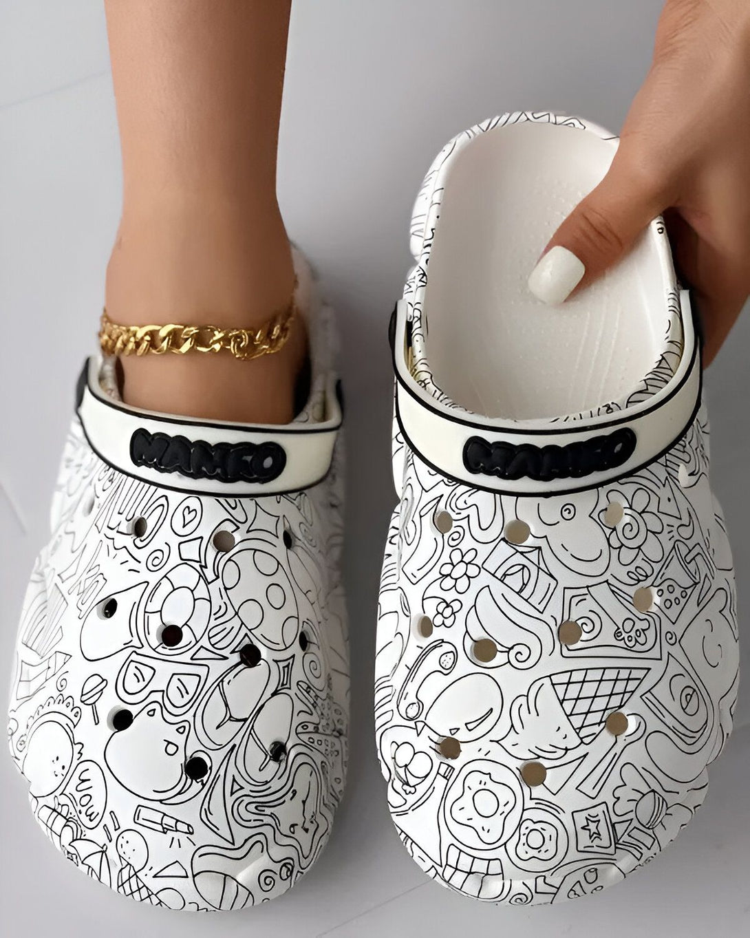Une paire de Crocs blancs avec des motifs de dessins animés noirs. Les motifs incluent des personnages, des animaux, des fleurs et de la nourriture.