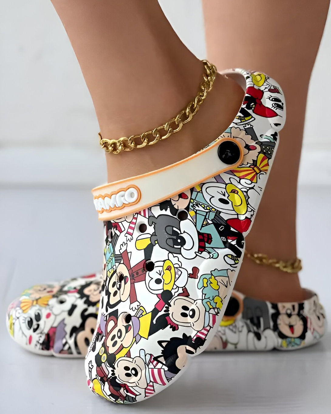 Une paire de sabots blancs avec des motifs de dessins animés colorés. Les sabots sont portés par une femme qui a une chaîne dorée autour de la cheville.
