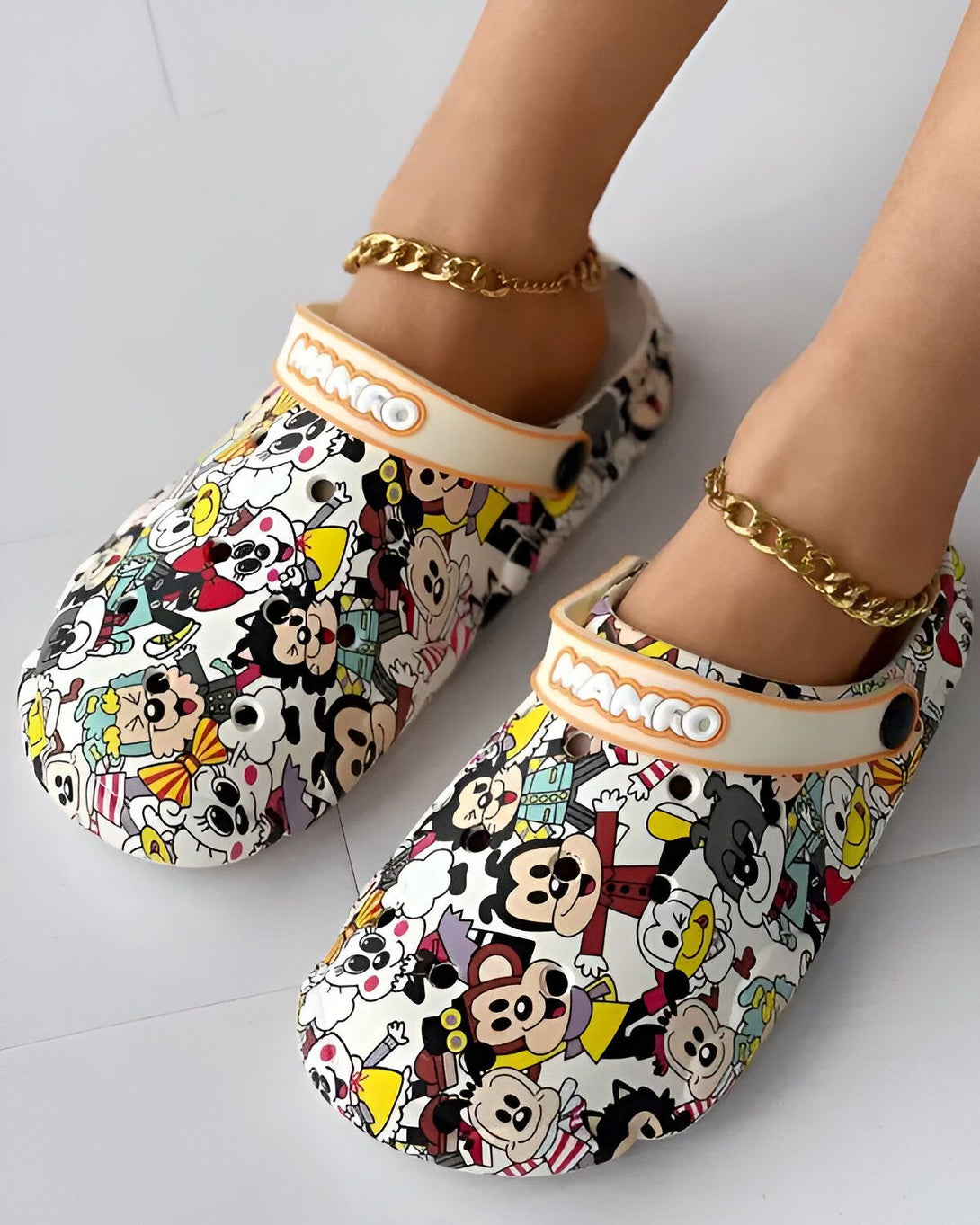 Une paire de sabots blancs avec des motifs de dessins animés colorés. Les sabots sont portés par une femme qui porte également une chaîne dorée autour de la cheville.