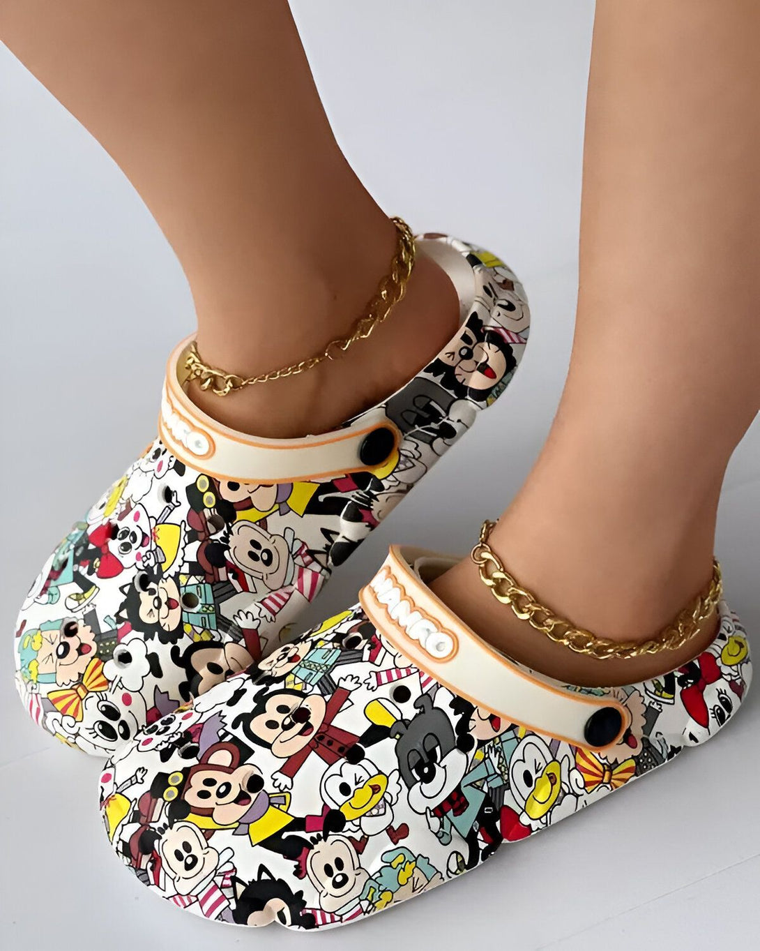 Une paire de sabots blancs à motifs de personnages de dessins animés Disney. Les sabots sont portés par une femme qui a des chaînes dorées aux chevilles.