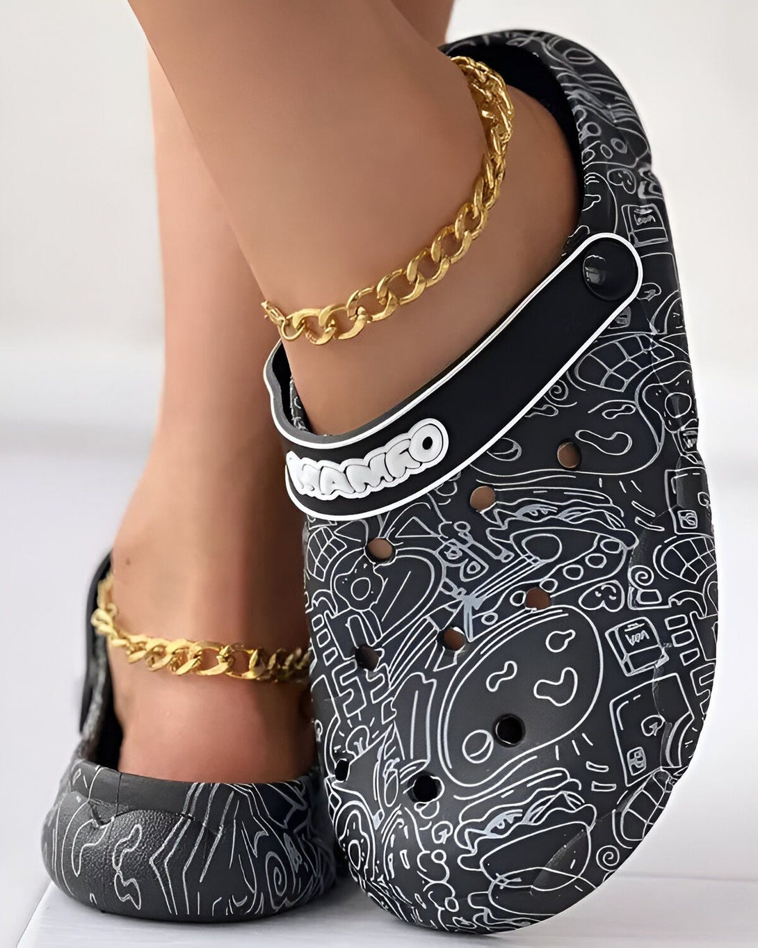 Une femme porte des sabots noirs et blancs avec des motifs de dessins animés. Elle porte également une chaîne de cheville en or.