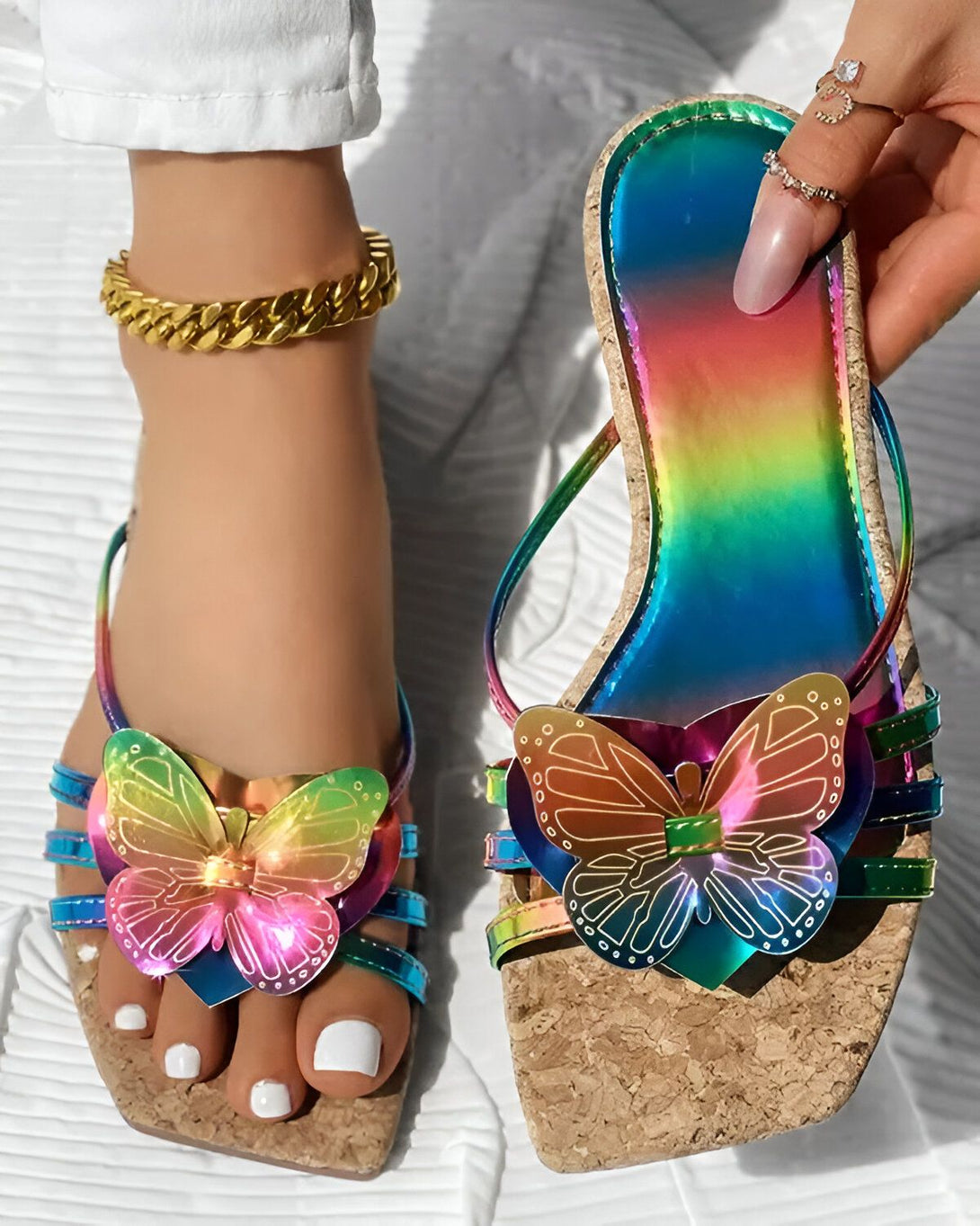Une femme porte des sandales plates à l'imprimé papillon. Les sandales sont multicolores et ont une semelle en liège. La femme porte également un pantalon blanc et un haut blanc. Elle a les ongles des pieds peints en blanc et porte un bracelet de cheville en or.