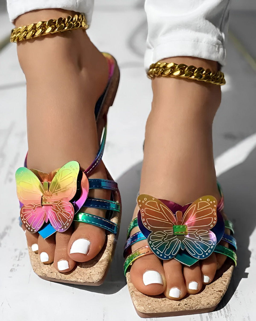Une femme porte des sandales plates à brides colorées avec un papillon multicolore sur le dessus. Elle porte également une chaîne dorée autour de la cheville.
