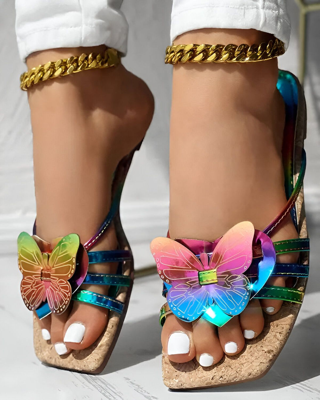 Une femme porte des sandales à talons compensés arc-en-ciel avec un papillon sur le devant. Elle porte également un pantalon blanc et un collier doré.