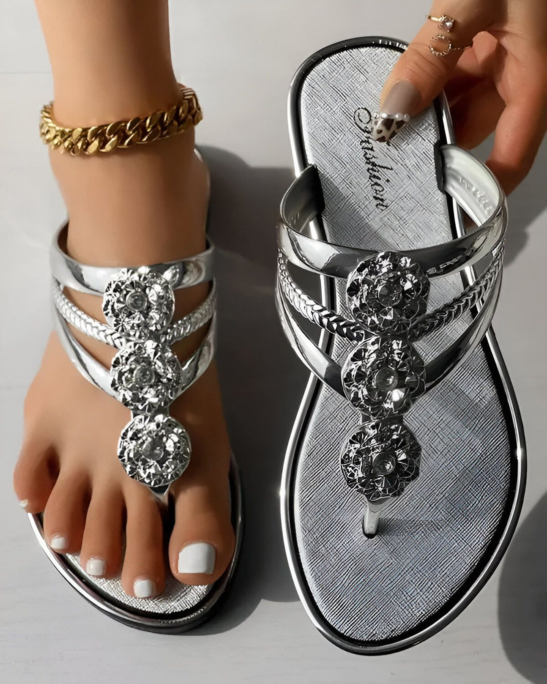 Une paire de tongs argentées avec des fleurs en strass. Les tongs sont portées par un pied droit avec une cheville fine et un bracelet de cheville doré. L'autre tong est tenue par une main avec des ongles manucurés.
