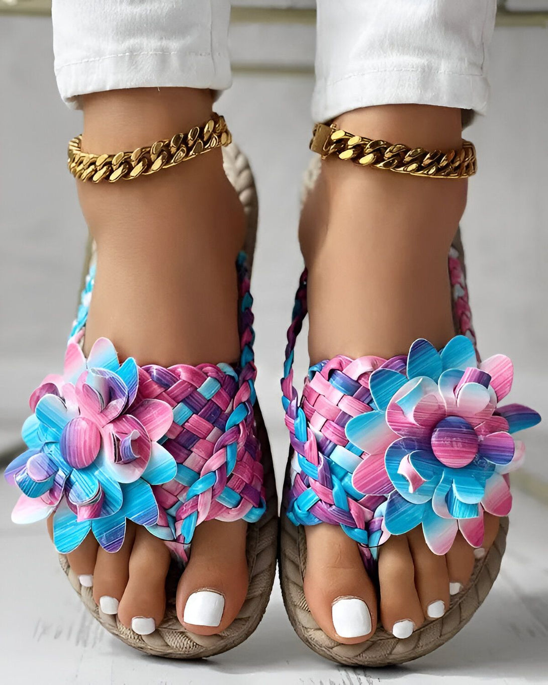 Une paire de sandales plates à fleurs pour femmes. Les sandales sont de couleur rose, bleue et blanche. Les fleurs sont de couleur rose, bleue et violette. Les sandales sont portées par une femme qui a les ongles des pieds peints en blanc. La femme porte également un pantalon blanc et des bijoux en or.