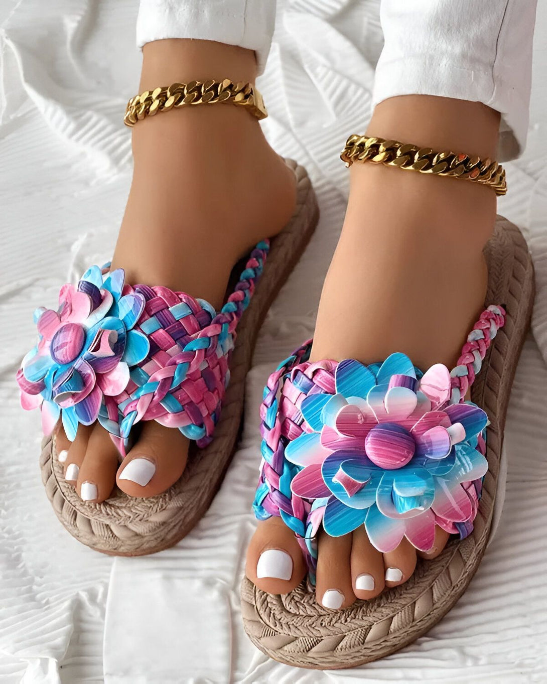Une paire de sandales plates avec des fleurs colorées sur le dessus. Les sandales sont portées par une femme avec un pantalon blanc et des chevilles dorées.