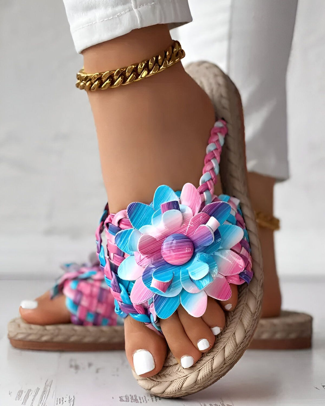 Une femme porte des sandales plates à fleurs colorées. Les sandales sont faites de cuir et ont une semelle en corde. La femme porte également un pantalon blanc et un bracelet de cheville doré.