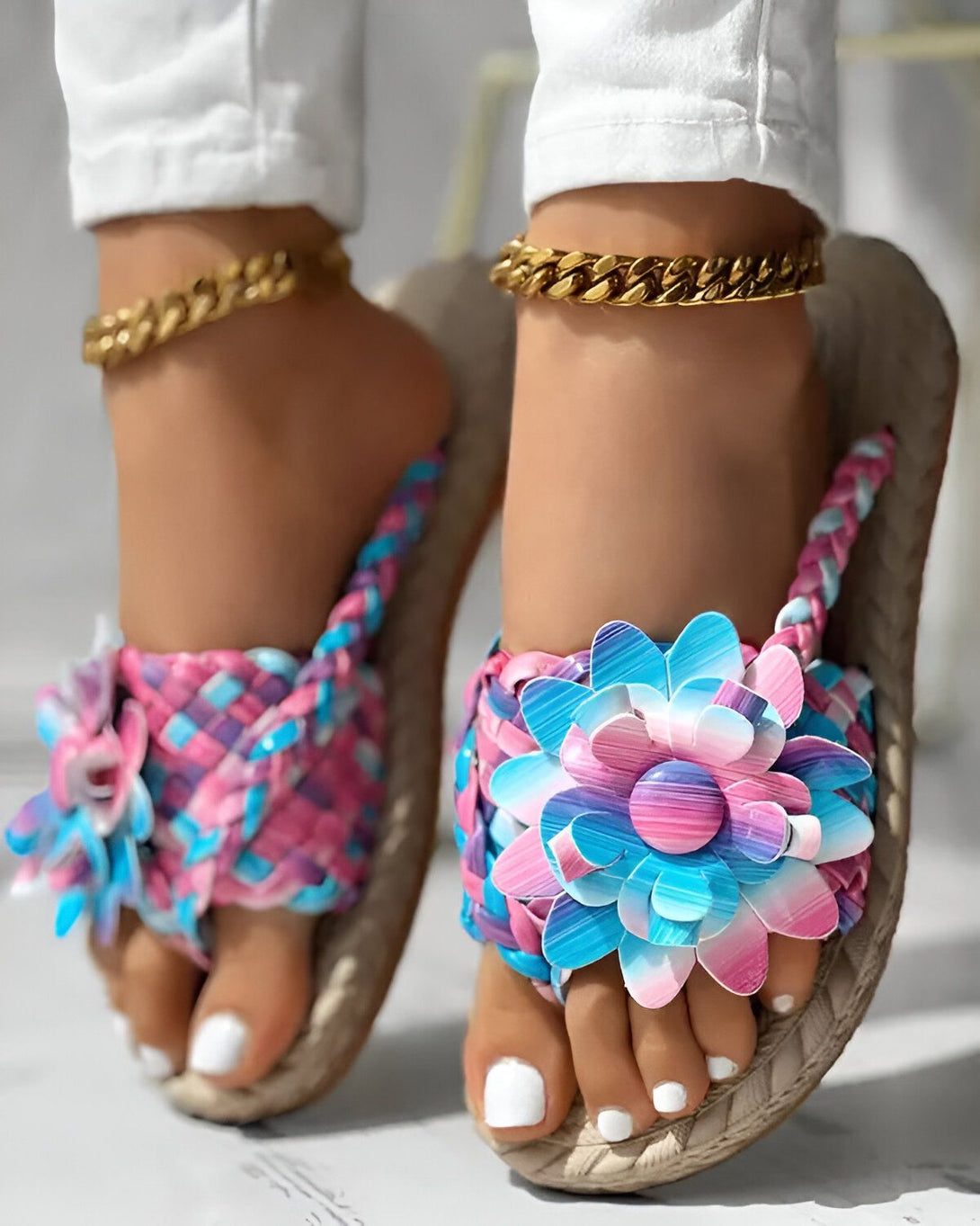 Une femme porte des sandales plates à fleurs colorées. Les sandales sont roses et bleues avec une fleur sur le dessus. La femme porte également un pantalon blanc et des bijoux dorés.