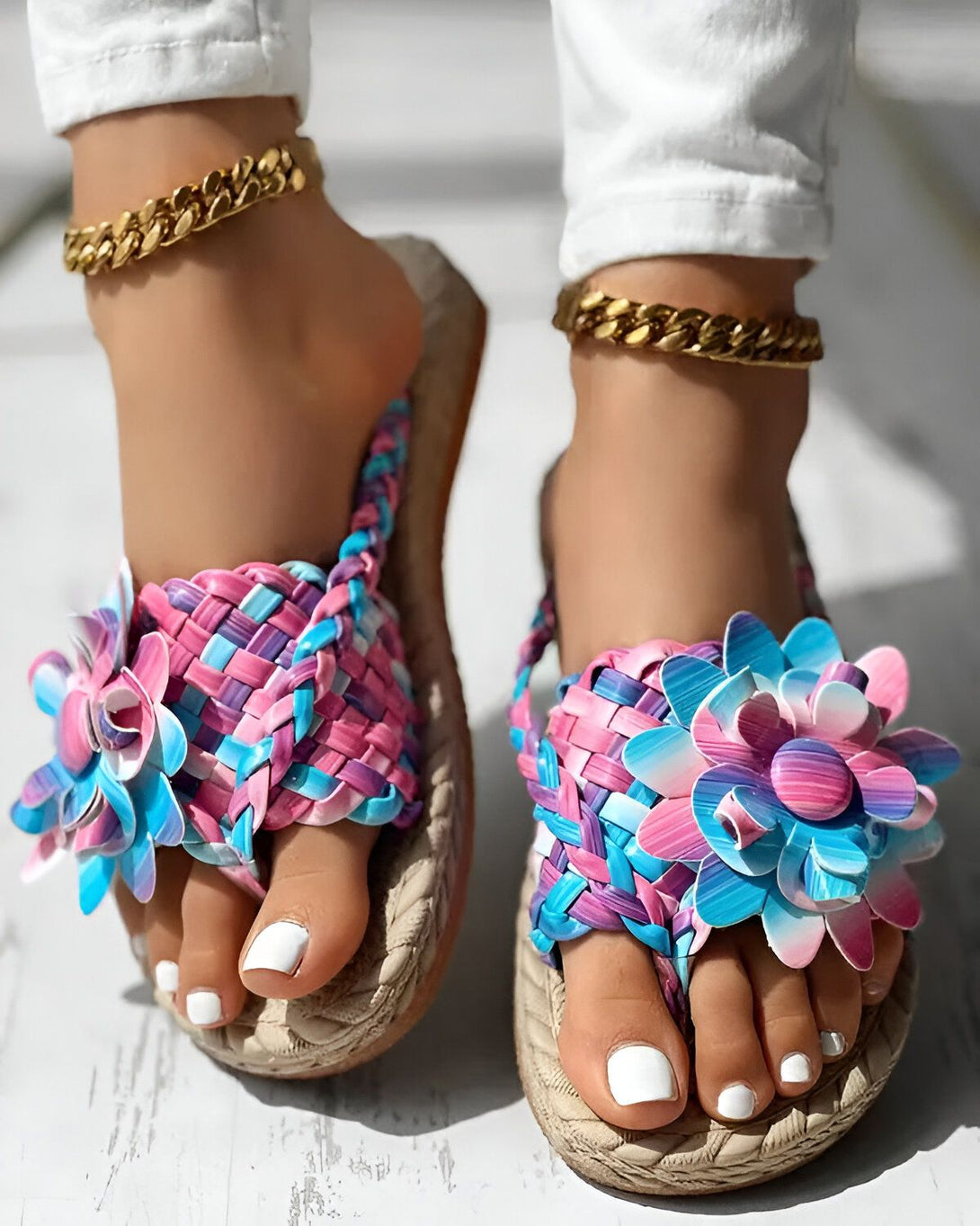 Une femme porte des tongs plates à fleurs colorées. Les tongs sont roses, bleues et violettes. La femme a les ongles des pieds peints en blanc et porte des bracelets de cheville dorés.